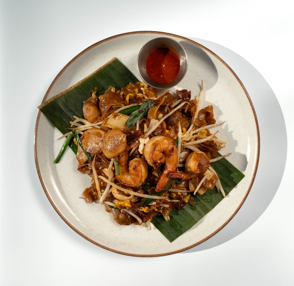 Char kway teow — wok-charred flat noodles with prawns, fish cake, bean sprouts, and chives on banana leaf